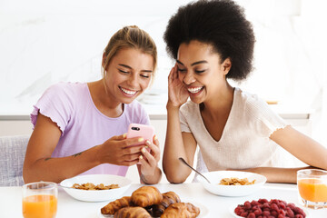 Nice cheerful multicultural girls using cellphone while having breakfast