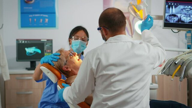 Senior woman sitting in dental clinic taking care of tooth health. Orthodontist lighting the lamp until examination, patient opening mouth lying on stomatological chair while nurse halping doctor.