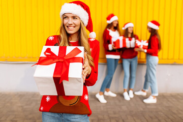Beautiful smiling young woman wearing santa claus hat with christmas present standing against background of happy friends celebrating christmas