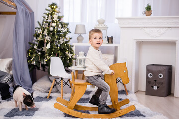 Happy Little blond boy in a white knitted sweater and jeans rides a wooden rocking horse in the children's room next to a Christmas tree with garlands. Cheerful childhood. Copy space, festive interior
