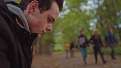 Young guy with a phone in his hands sitting on a bench in the park. In the background, a group of friends walking in the park. The concept of loneliness and dependence on the Internet and smartphones.