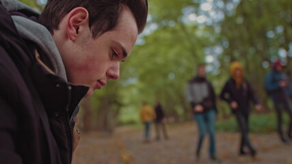 Young guy with a phone in his hands sitting on a bench in the park. In the background, a group of friends walking in the park. The concept of loneliness and dependence on the Internet and smartphones.