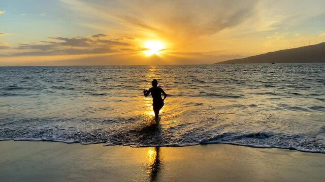 Silhouette of woman in a dress getting wet standing in Hawaii shoreline water taking a photo with her cell phone at sunset. Hazy golden sun with part of island in the distance across calm ocean.