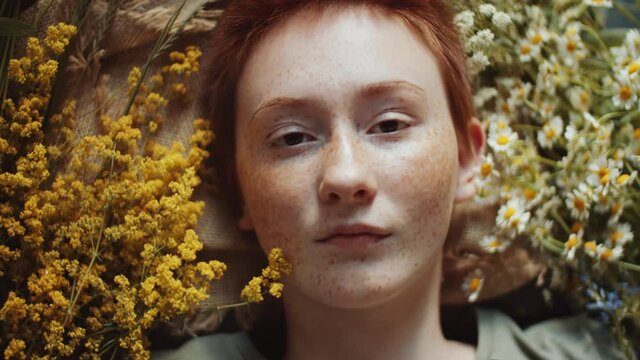 Top down shot of young beautiful woman with short ginger hair and freckles lying with eyes closed, on burlap fabric among wildflowers, then opening eyes and looking at camera