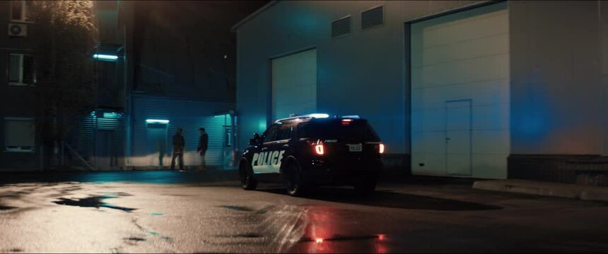 WIDE Police car turns on flashing lights while approaching two multi-ethnic males suspects near industrial buildings at night. Shot on RED cinema camera with 2x Anamorphic lens