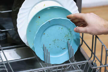 A man loads dirty dishes, plates, spoons, forks, cutlery into dishwasher tray.