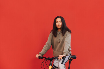 Fototapeta na wymiar Stylish lady in sports casual clothes rides a bicycle on a red background and poses for the camera. Woman walking on a bicycle, isolated. Isolated.