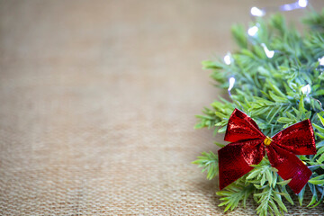 close-up of a red shiny and glossy bow with wreath and white lights, christmas decoration with ribbon and green leaves on jute, background, front view