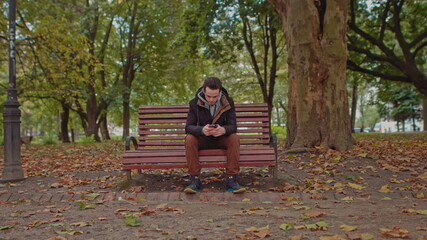 Young guy sitting on a park bench with a phone in his hands.Green tree young man stylishly dressed in the center of the frame. The guy uses a smartphone