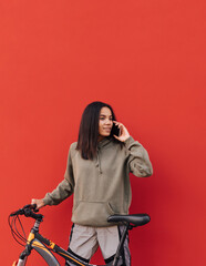 Naklejka na ściany i meble Portrait of a cute hispanic woman in sportswear standing with a bicycle on a background of a red wall and calling on the phone with a smile on his face. Vertical.