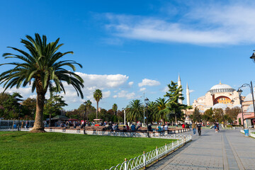 Obraz premium The central square of Istanbul. Palm trees and a crowd of tourists. Hagia Sophia in the summer morning. Red bench for the sale of simit.