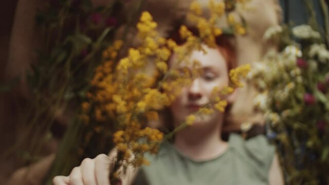 Top down rack focus shot of young charming woman with short ginger hair and freckles lying on burlap fabric among wildflowers and holding yellow meadow flower close to camera