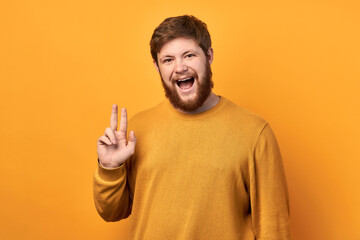 Portrait of a cheerful man with a beard showing the sign of the horns, enjoying a rock and roll party in a yellow sweater, isolated on yellow background. shows 2 fingers