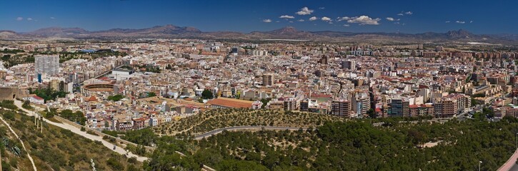 Obraz premium A viewpoint from a hill on the Spanish city Alicante and the mountains against blue sky. A panoramic view of a valley with green trees and roads in the forefront.