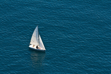 Aerial view on a white floating sailboat with two people in it in the blue Mediterranean sea.
