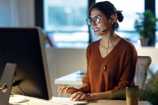 Beautiful young business woman working with computer while talking with earphone sitting in the office.