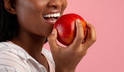 Dental care. Cropped view of black lady with healthy teeth eating ripe red apple over pink background, copy space