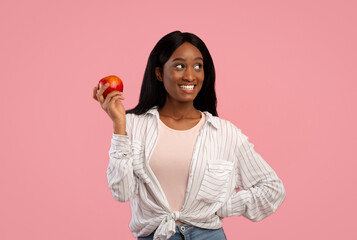 Portrait of lovely young black woman holding ripe red apple on pink studio background