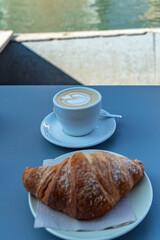 Cup of coffee and croissant in italian coffee bar in Venezia, Venice, Italy. Cappuccino and cornetto on plastic table.