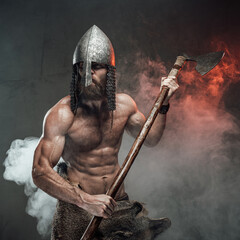 Naklejka premium Furious and shirtless viking with helmet on him and muscular build going to battle holding two handed axe in smoky background.
