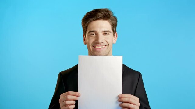 Portrait of young handsome businessman in suit holding white vertical a4 paper isolated on blue studio background.Copy space.
