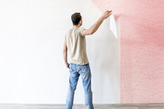young man tearing old paperhangings off the wall during renovation