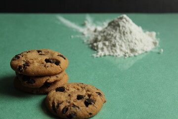 two or more cookies with chocolate drops are lying next to each other and a hill on a green grass background behind in the background is a hill of flour or powdered sugar there is a copy space