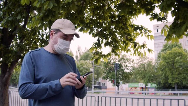 Adult caucasian 40 years old man wearing protective mask and casual clothes walking on a city street, using wireless internet on a smart phone. E-commerce at coronavirus pandemic time. Slow motion.