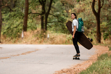 Fototapeta premium young sportive woman riding skateboard with wakeboard in her hand and backpack on her shoulders.