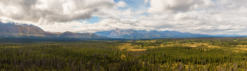Beautiful Panoramic View of Lush Forests, Trees and Land surrounded by Mountains on a Cloudy Summer Day in Canadian Nature. Aerial Drone Shot. Taken near Alaska Highway, Yukon, Canada.
