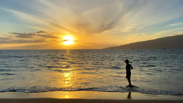 Medium shot of the silhouette of a woman in a dress standing in the ocean water at the Hawaii shore at sunset taking a picture with her cell phone with an island in the distance across lots of ocean
