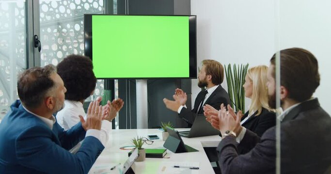 Close up of good-looking happy experienced mixed race businesspeople are satisfied of results of joint work and clapping hands simultaneously looking on big green screen chroma mock-up