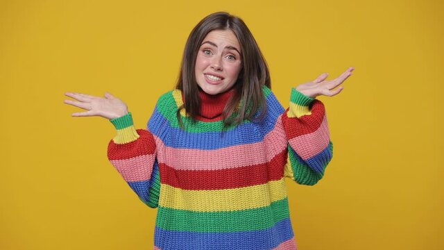 Confused puzzled young woman 20s years old in colorful sweater posing isolated on yellow background studio. People lifestyle concept. Looking camera spreading hands say oops biting lips gnawing nails
