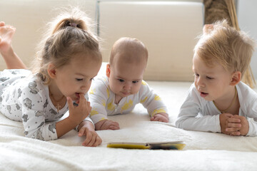 Three Little smiling kids play together on soft white bed at home. Brother and siste read book fairy tales with baby. Children meeting new born sibling.Toddlers laugh and bond. funny.Copy space