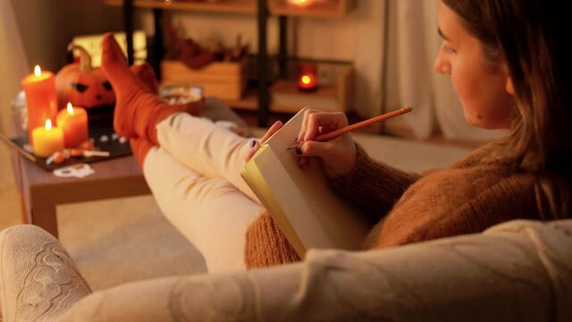 halloween, holidays and leisure concept - young woman with pencil writing to diary and resting her feet on table at cozy home