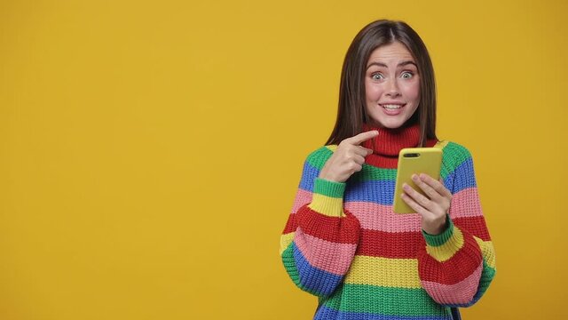Surprised young woman 20s years old in colorful sweater isolated on yellow background studio. People lifestyle concept. Using mobile cell phone typing sms mesage pointing finger aside showing thumb up