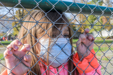 Little girl child in medical mask behind an iron fence