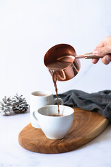 Two white cups with hot chocolate or cocoa drink on a wooden board on a light marble background with festive winter decorations. Female hand pouring drink into a cup. Winter mood. Selective focus 