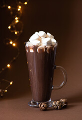 Cocoa drink with marshmallows in a glass on a brown background with festive winter decorations. Winter mood. Minimalistic monochrome composition. Selective focus. Frontal view. Vertical. Copy space