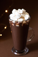 Cocoa drink with marshmallows in a glass on a brown background with festive winter decorations. Winter mood. Minimalistic monochrome composition. Selective focus. Frontal view. Vertical. Copy space