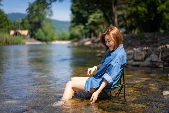 Smiling short haired young Asian girl in denim shirt and short pants sitting on camping chair in the clear stream with her feet in the stream and touching water with soft-focus of woods in background.