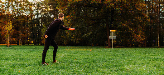 Young caucasian man playing disc golf on autumn play course with basket - 388334540