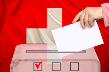 Obraz premium female voter lowers the ballot in a transparent ballot box on the background of the national flag of Switzerland, concept of state elections, referendum