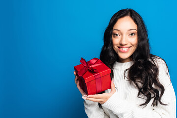 cheerful brunette woman in knitted sweater holding christmas present isolated on blue