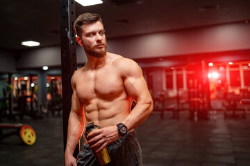 Fototapeta na wymiar Portrait of athletic well built man with six pack abs. Half turned to the camera. Gym background. Guy holding bottle. Closeup.