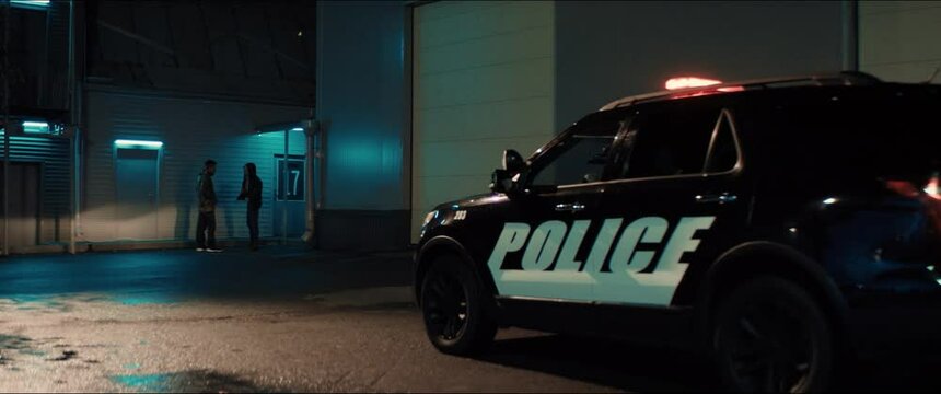 WIDE Police car turns on flashing lights while approaching two multi-ethnic males suspects near industrial buildings at night. Shot on RED cinema camera with 2x Anamorphic lens
