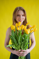 Caucasian woman with an armful of yellow tulips on a yellow background. International Women's Day. Bouquet of spring flowers