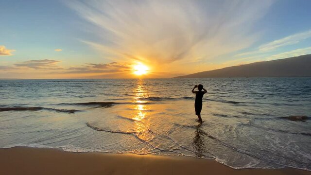 Silhouette of a woman in a dress standing in the gentle surf of an Hawaii beach at sunset taking a photo with her cell phone of the island in the distance and the reflection of the sun on the ocean