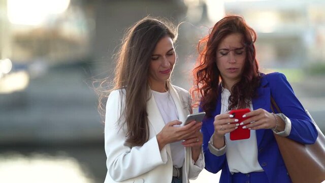 two women are viewing photos in smartphones, standing outdoor in city at sunny day, portrait of townswomen