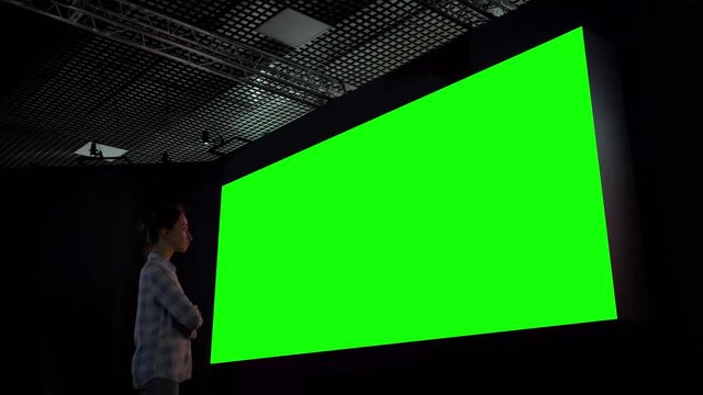 Woman looking at large wall blank interactive green display in dark room of modern technology exhibition or museum. Mock up, green screen, futuristic, template, education, chroma key concept